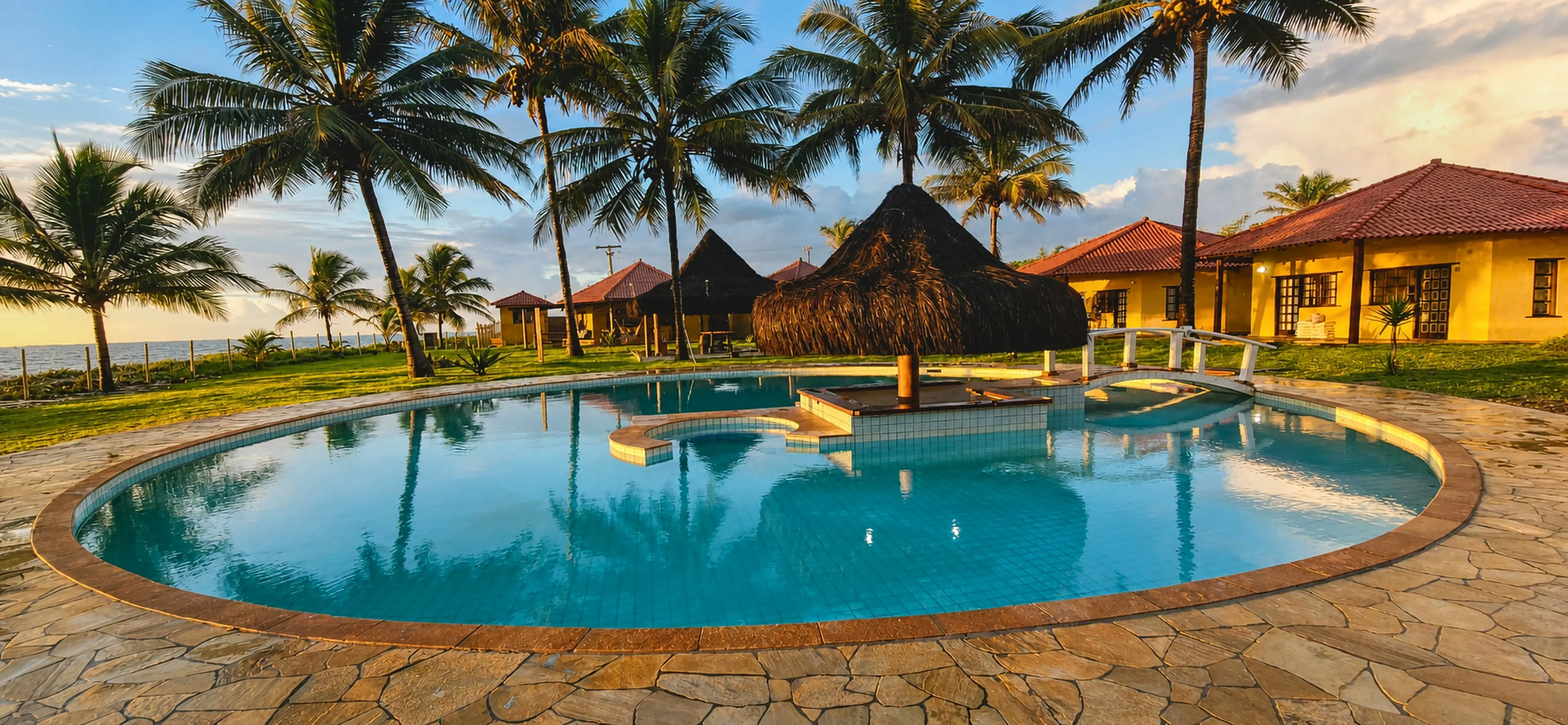 Área do Condomínio Praia dos Corais: piscina diante dos bangalôs e dos coqueiros, ao pôr do sol